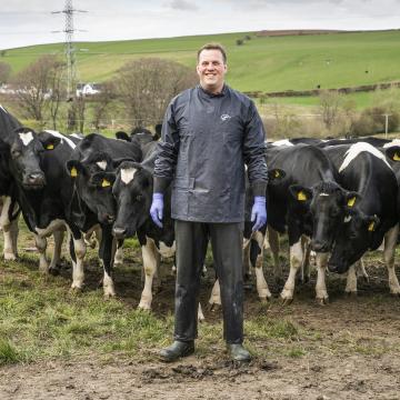 A photograph of a farmer stood in front of a herd of cows